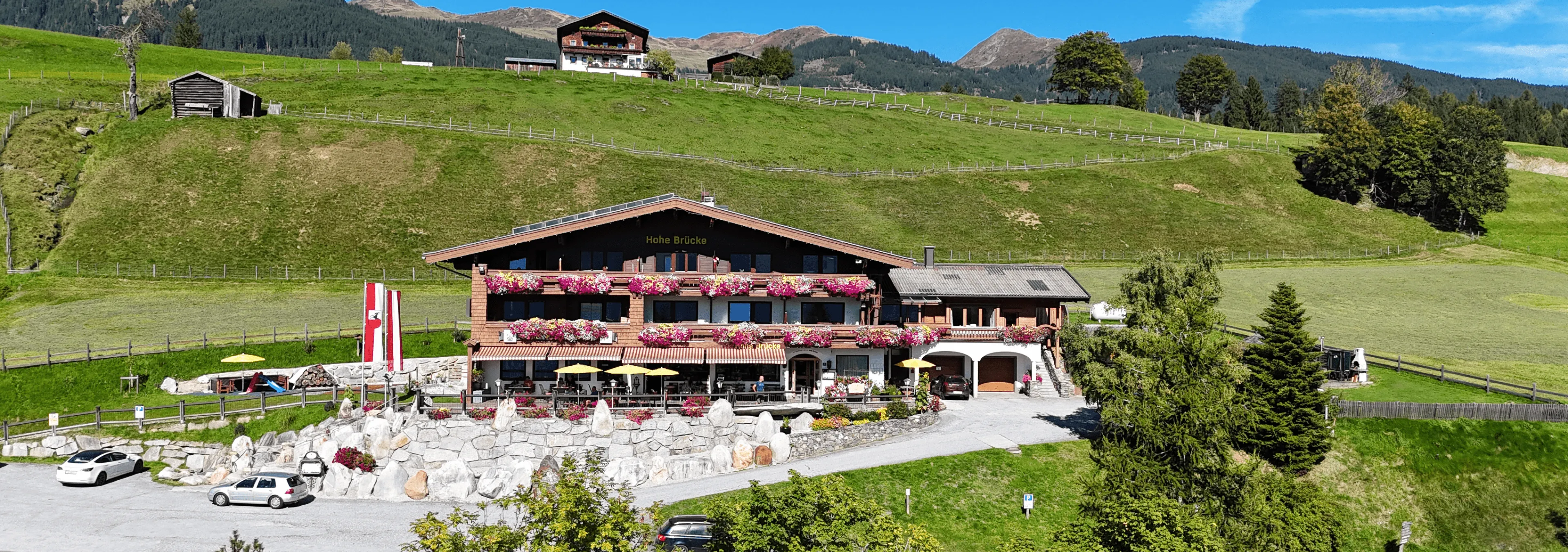 Landschaft mit einem Gasthaus, umgeben von Hügeln und Wiesen, im Hintergrund das Gebirge.